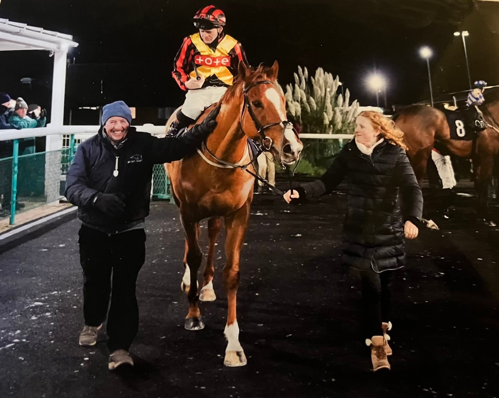A chestnut racehorse returning from a night race with the team gathered around