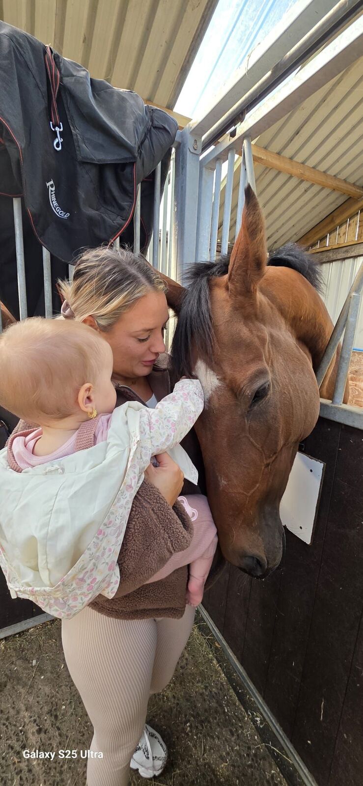 A woman holds a baby as the baby gently touches a horse’s head at the stable door.